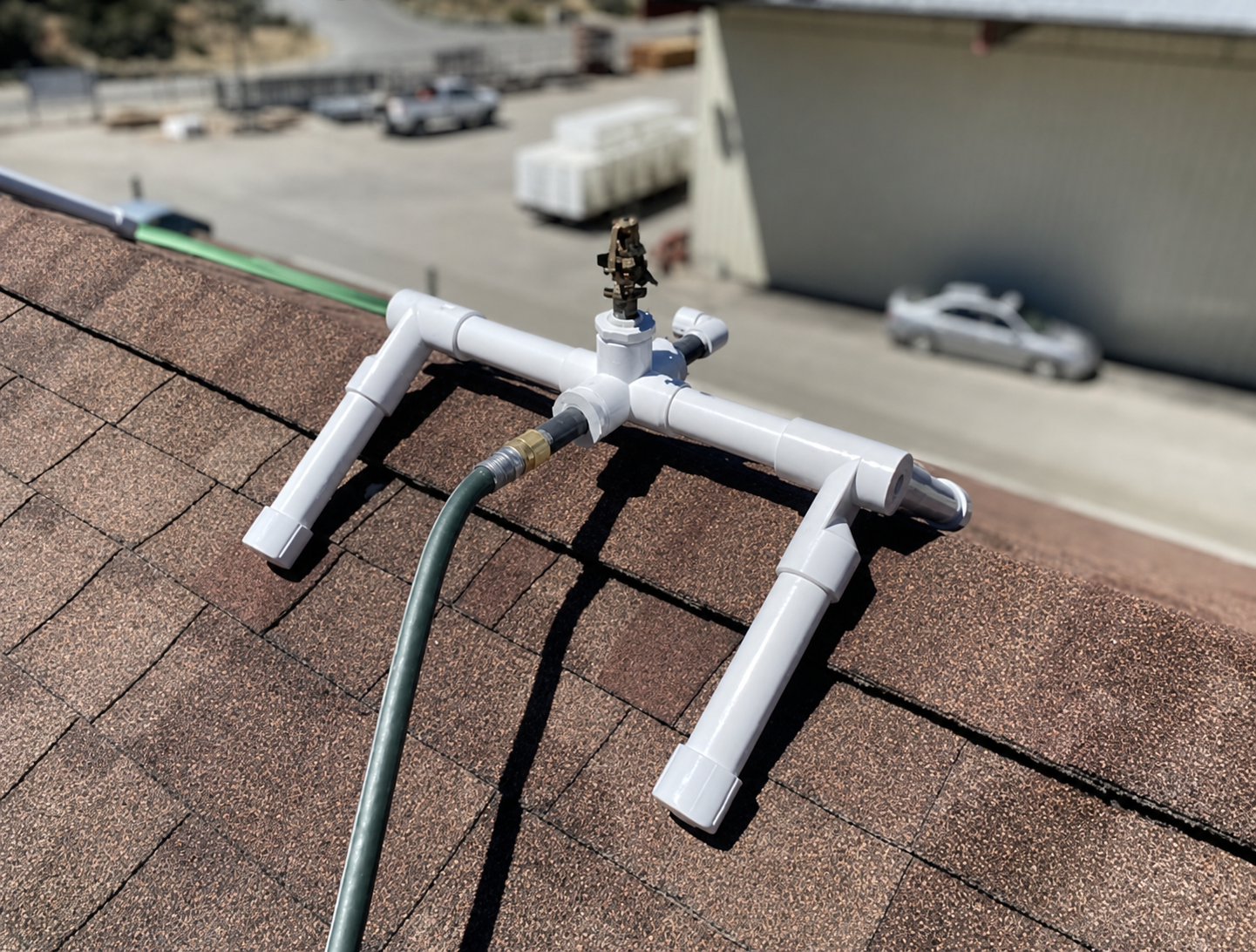 Inexpensive PVC pipe roof sprinkler assembly sitting on an asphalt shingle ridge, fed by a garden hose